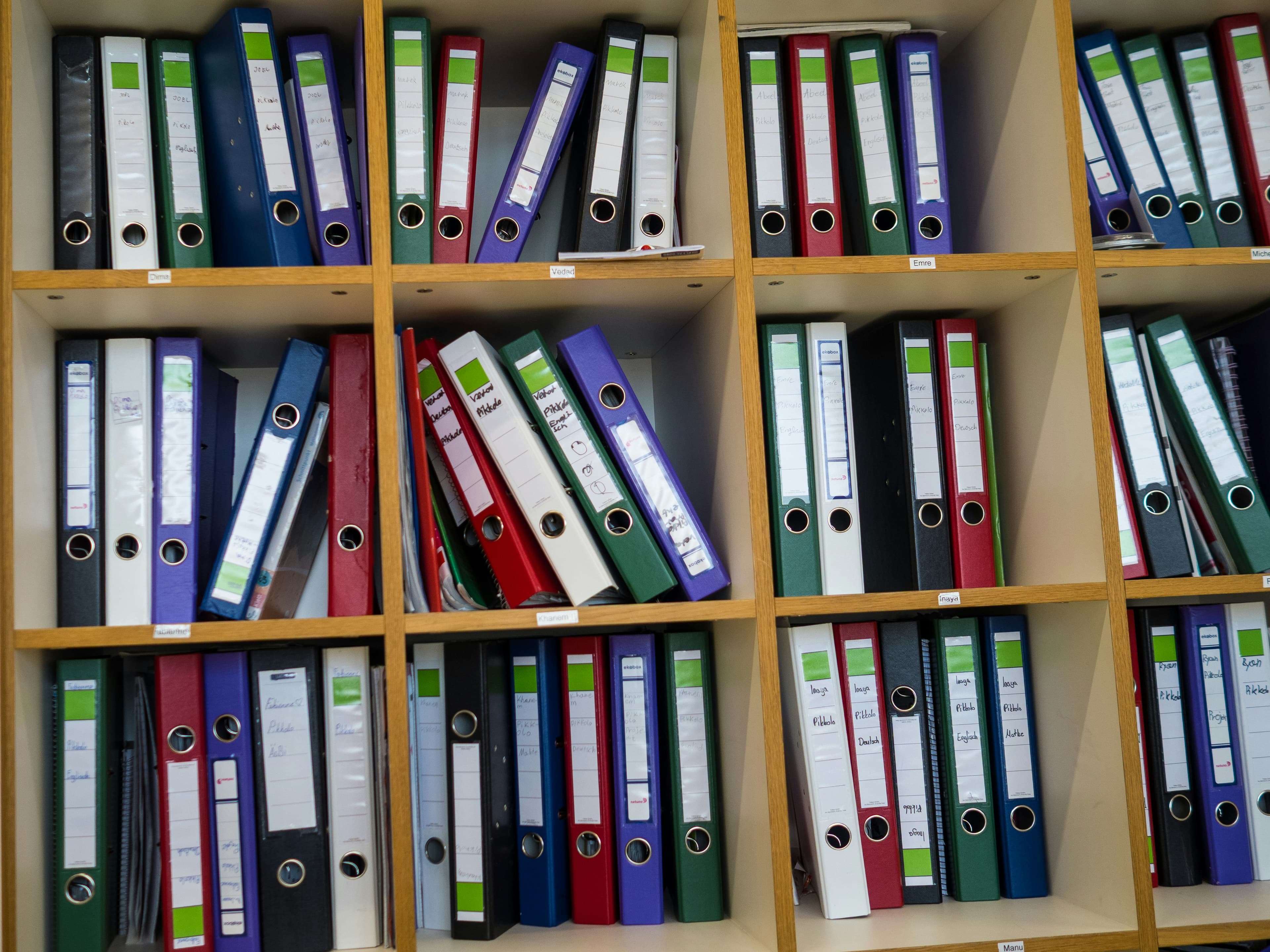 An overstuffed wooden shelf of color-coded ring binders, some leaning and crowded out.
