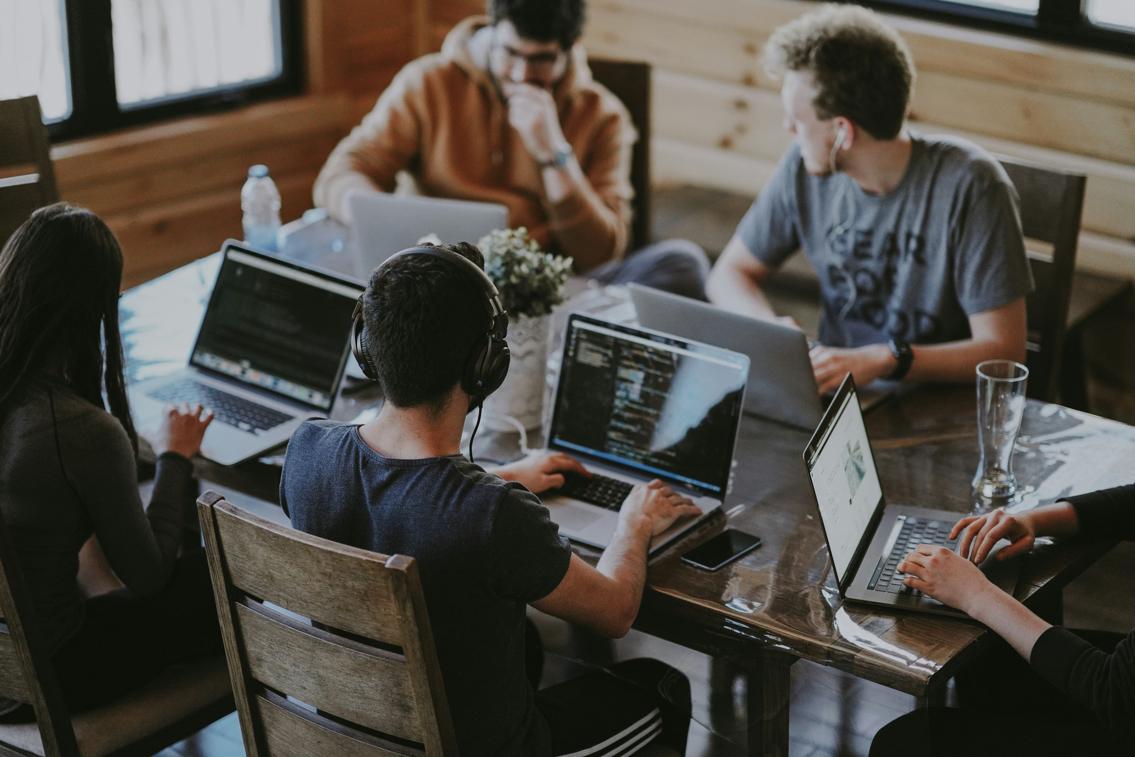 Five people hunched over laptops at a shared wooden table, a small plant in the middle.