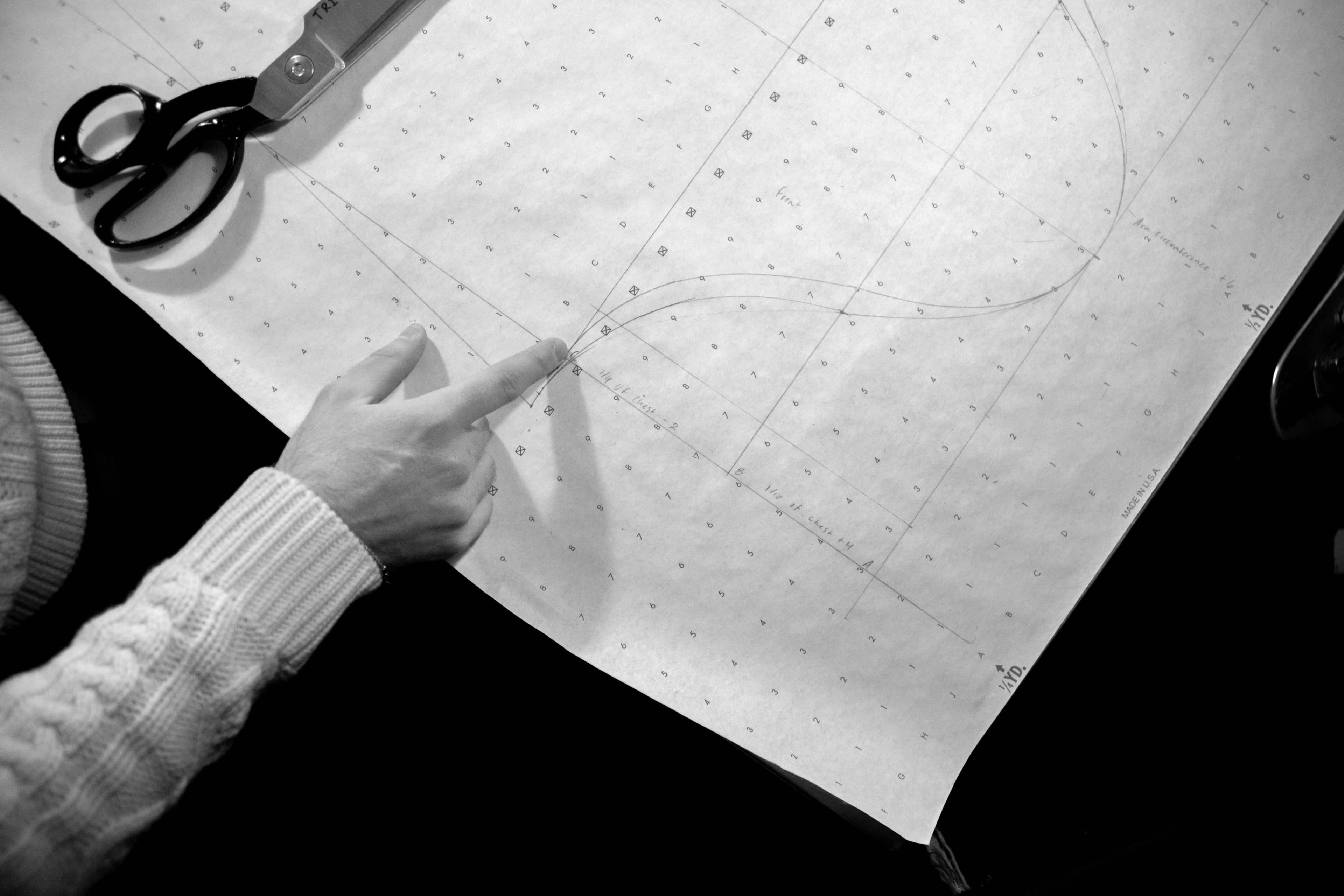 An overhead black-and-white shot of hands drafting a garment pattern on gridded paper, dressmaker's shears resting at the edge.